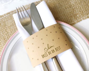 May include: A place setting with a white plate featuring a pink and gray rim, a folded white napkin, and silverware. A brown paper band with floral designs and the text "Schön, DASS DU DA BIST" wraps the napkin and silverware.