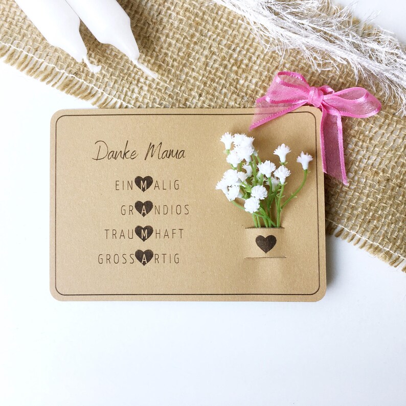 May include: A brown card with the text "Danke Mama" and the words "einmalig", "grandios", "traumhaft", and "grossartig" with a heart replacing the "M" in each word. A small bouquet of white flowers is in a brown heart-shaped vase.