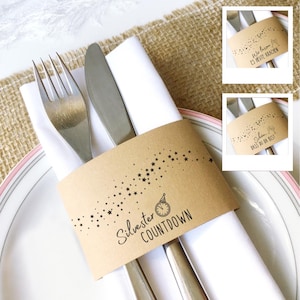 May include: A festive table setting with silverware and a white napkin, held together by a brown paper band. The band is decorated with black stars and the words "Silvester Countdown" and a clock illustration.