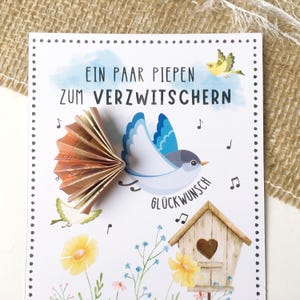 May include: A white card with a blue bird holding money in its beak. The card has the text "Ein paar piepen zum verzwitchern" and "Gl&uuml;ckwunsch" on it. The card is decorated with flowers and a birdhouse.