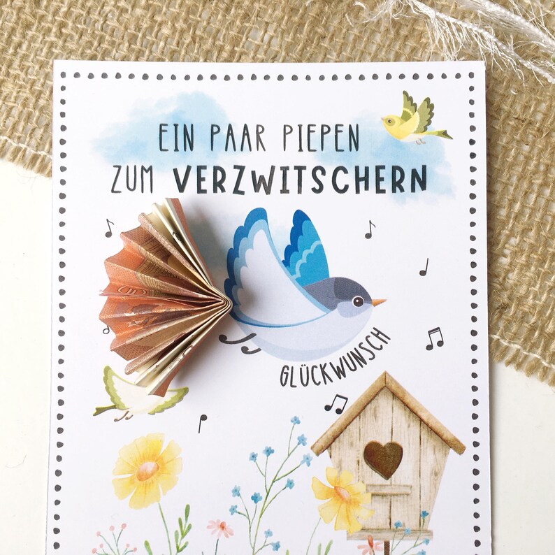 May include: A white card with a blue bird holding money in its beak. The card has the text "Ein paar piepen zum verzwitchern" and "Gl&uuml;ckwunsch" on it. The card is decorated with flowers and a birdhouse.
