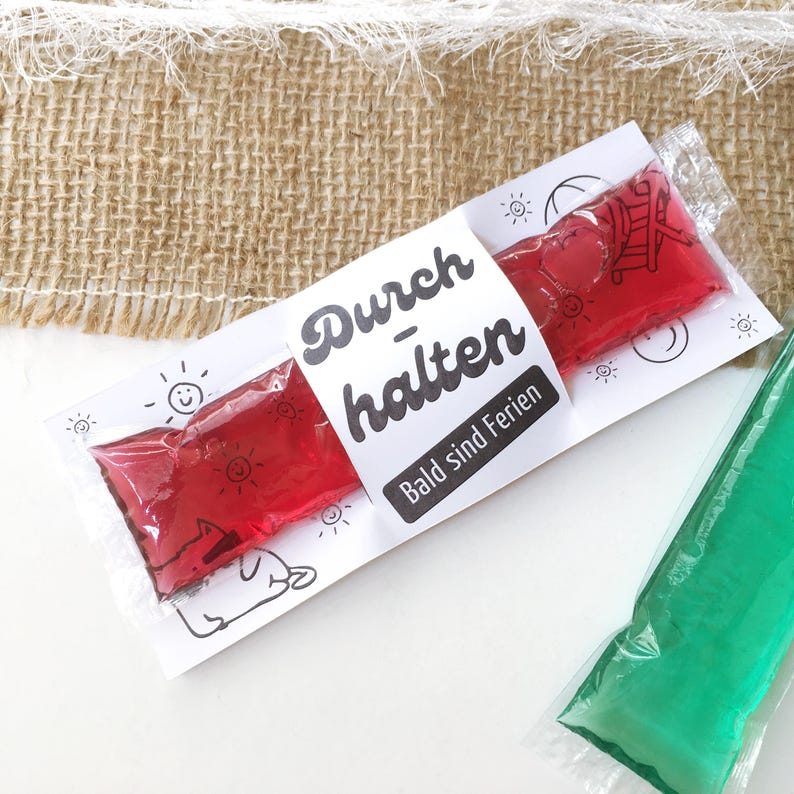 May include: A package containing a red, heart-shaped frozen treat, with the German text "Durchhalten Bald sind Ferien" on a white label. The label is decorated with sun and dog illustrations. A green frozen treat is visible in the background.