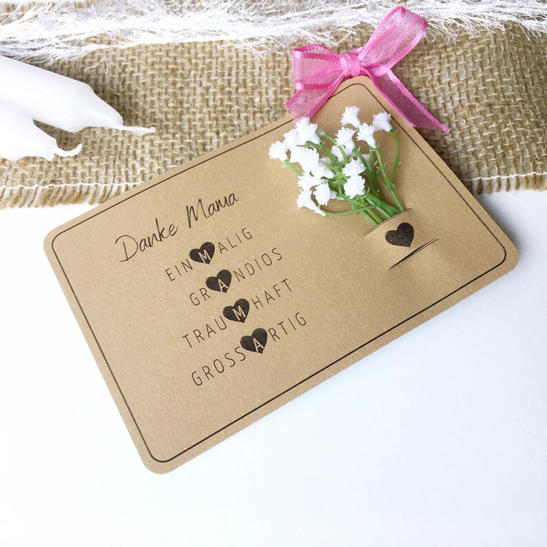 May include: A brown card with the text "Danke Mama" and the words "einmalig, grandios, traumhaft, grossartig" with a heart next to each word. The card has a small bouquet of white flowers with a brown heart on the side.