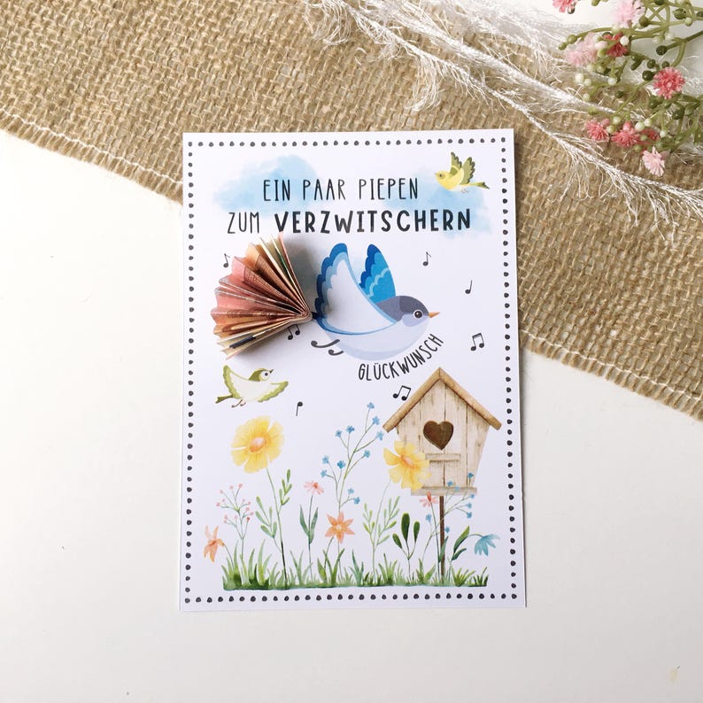 Geldgeschenkkarte zum Geburtstag. Farbenfrohe Glückwunschkarte mit Vögeln, Musiknoten, Blumenwiese und Vogelhäuschen im Hintergrund. Zusammengefalteter Geldschein in Fächerform ist als Flügel aufgeklebt. Rundum Punkte. Druckvorlage zum selbstausdrucken.