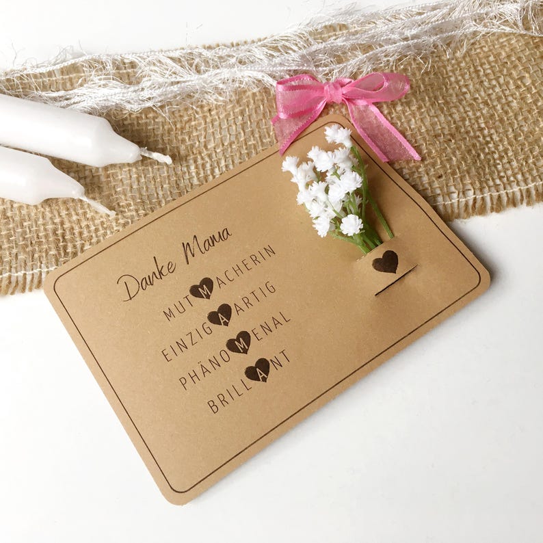 May include: A brown card with the text "Danke Mama" and a list of words with hearts: "Mutmacherin", "Einzigartig", "Ph&auml;nomenal", "Brilliant". A small bouquet of white flowers is attached to the card with a pink ribbon.