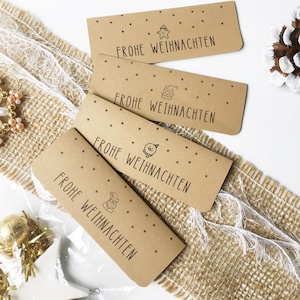 Könnte beinhalten: Vier rechteckige Geschenk-Anhänger aus Kraftpapier mit dem Text "FROHE WEIHNACHTEN" und Stern- und Weihnachtsillustrationen. Die Anhänger liegen auf einer Jutefläche.