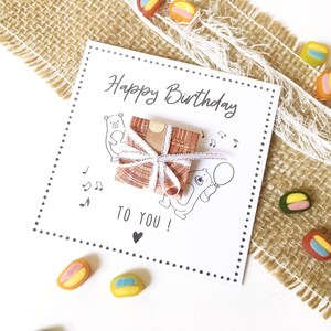 May include: A white birthday card with the text "Happy Birthday TO YOU!" and illustrations of bears and musical notes. A small gift wrapped in brown paper and tied with white ribbon sits on the card. Colorful candies are scattered around the card.