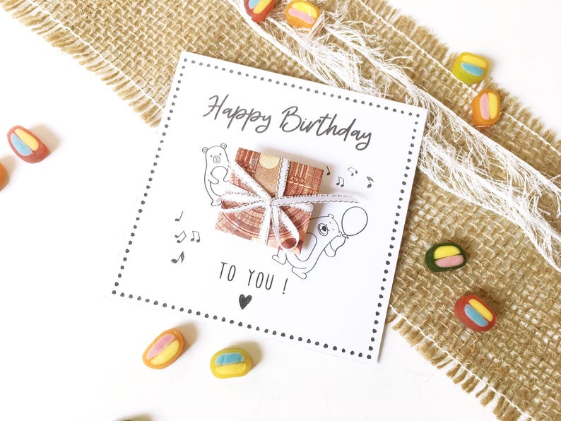 May include: A white birthday card with the text "Happy Birthday TO YOU!" and illustrations of bears and musical notes. A small gift wrapped in brown paper and tied with white ribbon sits on the card. Colorful candies are scattered around the card.