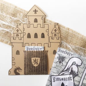 Könnte beinhalten: Einladung in Form einer Burg aus Pappe mit einer Drachensilhouette, Fleur-de-Lis und dem Wort "Burgpost". Enthält einen passenden Umschlag mit Wappen und eine gedruckte Einladung mit einem Ritterhelm.