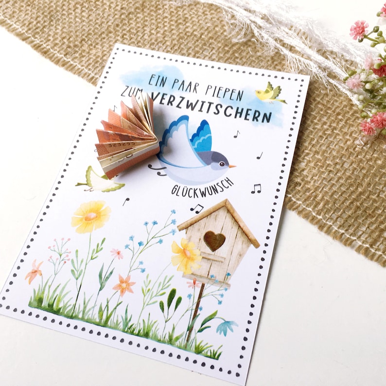 May include: A white card with a blue bird flying with a money fan in its beak. The card has the text "Ein paar piepen zum verzwitchern" and "Gl&uuml;ckwunsch" with musical notes. The card has a watercolor background with flowers and a birdhouse.
