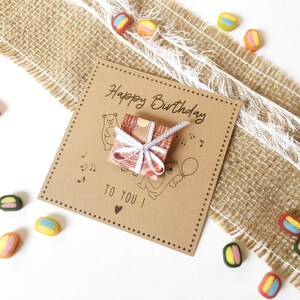 May include: A brown birthday card with the text "Happy Birthday TO YOU!" and illustrations of bears, musical notes, and a balloon. A small gift wrapped in patterned paper and tied with a white ribbon sits on the card. Colorful candies are scattered around.