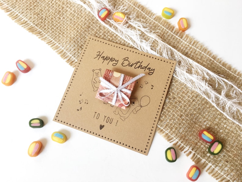 May include: A brown birthday card with the text "Happy Birthday TO YOU!" and illustrations of bears, musical notes, and a balloon. A small gift wrapped in patterned paper and tied with a white ribbon sits on the card. Colorful candies are scattered around.