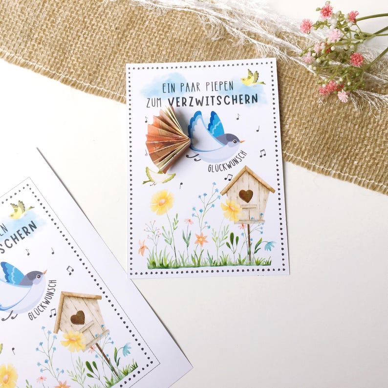 May include: A white card with a blue bird holding money in its beak. The card has the text "Ein paar piepen zum verzwitchern" and "Gl&uuml;ckwunsch" on it. The bird is flying over a small birdhouse with a heart-shaped opening.
