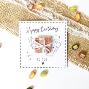 May include: A white birthday card with the text "Happy Birthday TO YOU!" and a gift illustration. The card is decorated with musical notes and a heart. Colorful candies are scattered around the card on a burlap and white background.