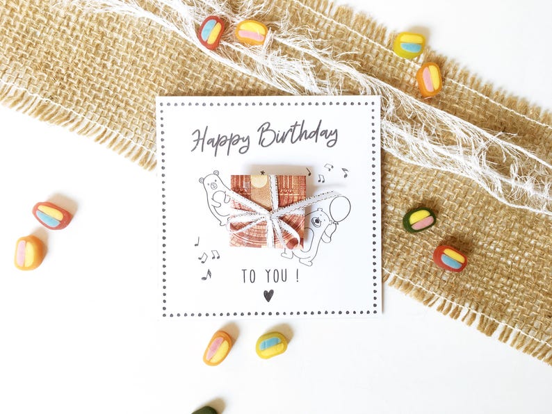 May include: A white birthday card with the text "Happy Birthday TO YOU!" and a gift illustration. The card is decorated with musical notes and a heart. Colorful candies are scattered around the card on a burlap and white background.