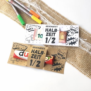 Puede incluir: Una tarjeta de papel kraft con las palabras "BRAVO!" y "GESCHAFFT HALB ZEIT 1/2" impresas, junto con un ratón de superhéroe de dibujos animados y la palabra "SUPER!". La tarjeta contiene una barra de chocolate. Se ven rotuladores de colores.