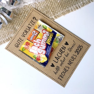 Pu&ograve; includere: Un biglietto marrone con il testo "GUTE VORS&Auml;TZE?" e "LACHEN hilft sofort bei Stress!" Il biglietto ha un piccolo pacchetto di caramelle gommose attaccato con il testo "Lach Gummi Minis" sulla confezione.