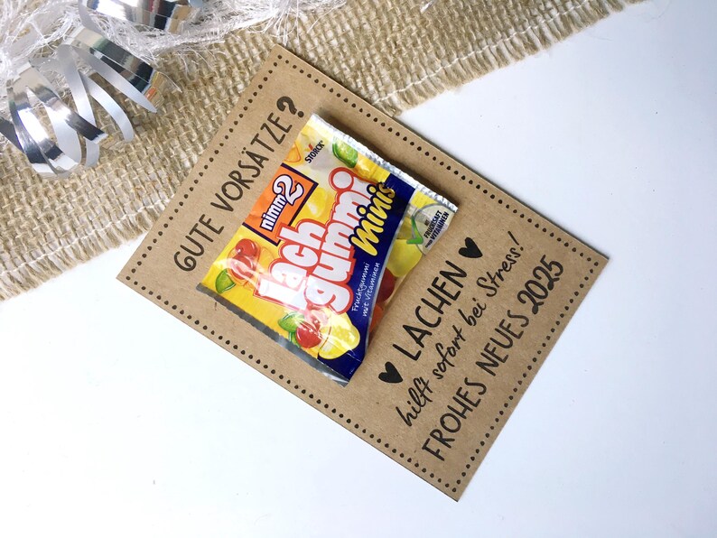 Pu&ograve; includere: Un biglietto marrone con il testo "GUTE VORS&Auml;TZE?" e "LACHEN hilft sofort bei Stress!" Il biglietto ha un piccolo pacchetto di caramelle gommose attaccato con il testo "Lach Gummi Minis" sulla confezione.