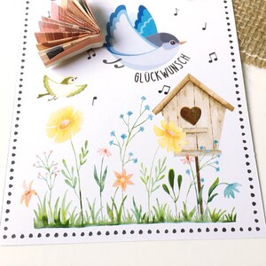 May include: A white card with a blue bird flying, a small yellow bird, a birdhouse with a heart cutout, and flowers. The card says "Gl&uuml;ckwunsch" which is German for congratulations.