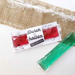 May include: Two colorful ice pops in clear plastic packaging. One is red, the other is green. A white label on the red ice pop reads "Durchhalten Bald sind Ferien" in black text, with small illustrations.