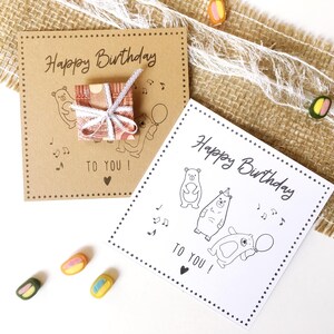 May include: Two birthday cards with the text "Happy Birthday TO YOU!" are displayed. One card is brown with a small gift box and bear illustrations. The other card is white with bear illustrations. Colorful candies are scattered around.