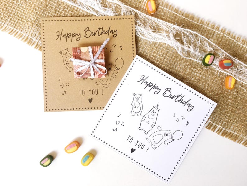 May include: Two birthday cards with the text "Happy Birthday TO YOU!" are displayed. One card is brown with a small gift box and bear illustrations. The other card is white with bear illustrations. Colorful candies are scattered around.