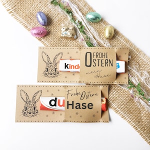 以下が含まれることがあります： ウサギのイラストと「Frohe Ostern mein Hase」の文字が入ったクラフト紙のイースターギフトホルダー2つ。各ホルダーにはチョコレートバーが入っています。アルミホイルで包まれたチョコレートエッグと小さなセラミックウサギが散らばっています。