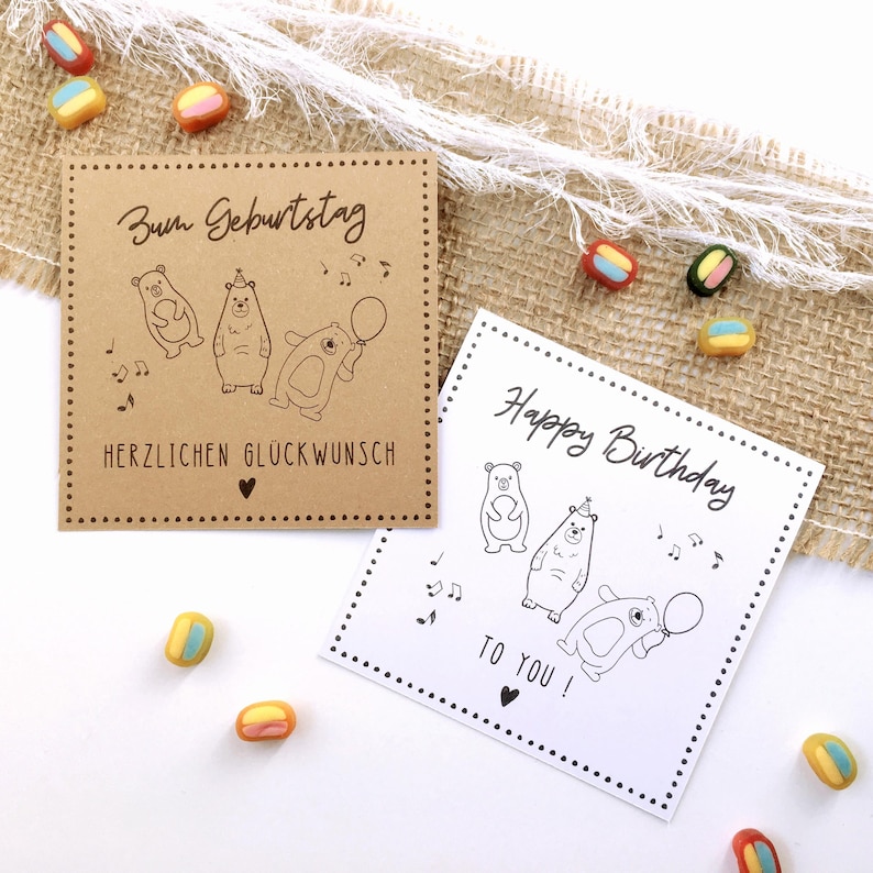 May include: Two birthday cards with bear illustrations. One card is brown with German text and the other is white with English text. Both cards have a dotted border and musical notes. Colorful candies are scattered around the cards.