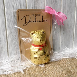 May include: A small chocolate teddy bear wrapped in gold foil with a red ribbon and a heart. The bear is sitting on a brown card with the text "Ein kleines Dankesch&ouml;n".