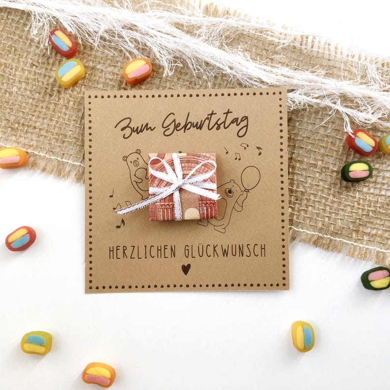 May include: A brown birthday card with the German text "Zum Geburtstag" and "Herzlichen Gl&uuml;ckwunsch". The card features illustrations of bears and musical notes, with a small gift tied with a white ribbon. Colorful candies are scattered around the card.