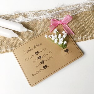 May include: A brown card with the text "Danke Mama" and a small bouquet of white flowers. The card has the words "Herzensmensch", "Alltagsengel", "Warmherzig", and "Wunderbar" with a heart symbol next to each word.