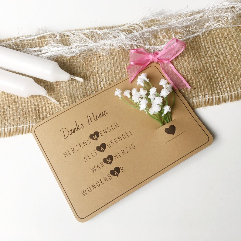 May include: A brown card with the text "Danke Mama" and a small bouquet of white flowers. The card has the words "Herzensmensch", "Alltagsengel", "Warmherzig", and "Wunderbar" with a heart symbol next to each word.