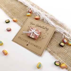 May include: A birthday card with the German text "Zum Geburtstag" and "Herzlichen Gl&uuml;ckwunsch" is shown. A small gift wrapped in patterned paper is tied with a white ribbon. Colorful candies are scattered around the card on a burlap and white surface.