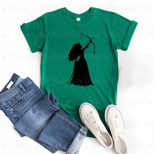Może przedstawiać: Zielony t-shirt z czarną sylwetką kobiety z długimi włosami trzymającej łuk i strzałę. Kobieta ma na sobie sukienkę.