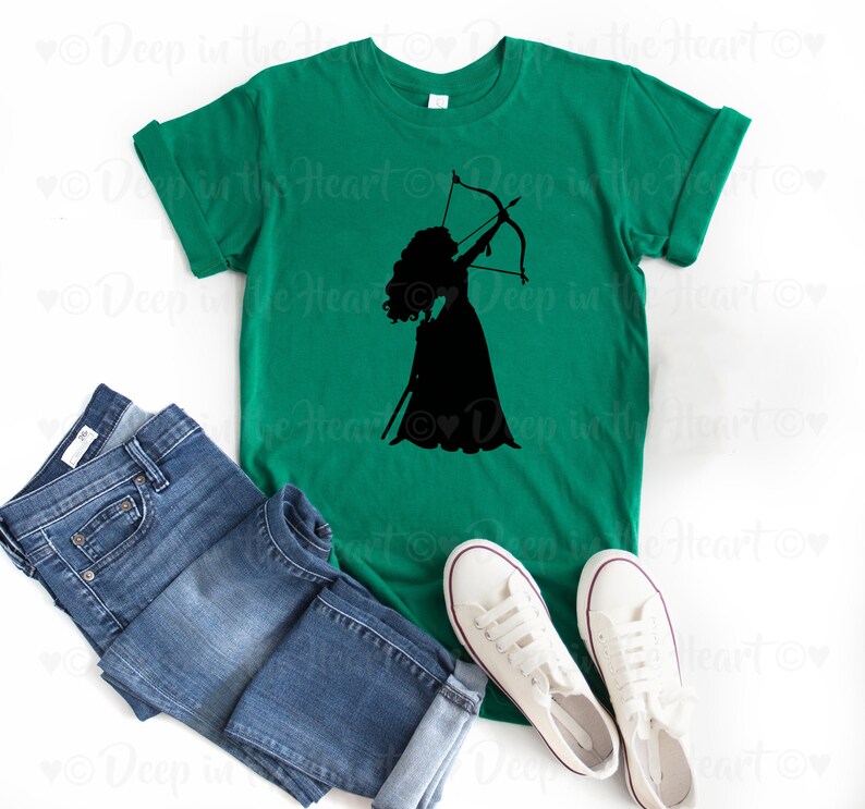 Może przedstawiać: Zielony t-shirt z czarną sylwetką kobiety z długimi włosami trzymającej łuk i strzałę. Kobieta ma na sobie sukienkę.