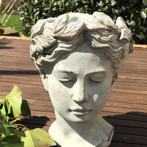 Könnte beinhalten: Eine verwitterte, grau getönte Steinkopfskulptur einer Frau mit geschlossenen Augen, geschmückt mit einem Lorbeerkranz. Die Skulptur steht auf einer Holzoberfläche, mit grünem Laub im Vordergrund. Ein dekoratives Gartenelement.