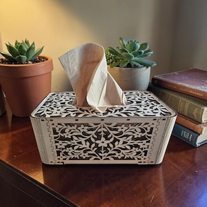 Peut inclure: Boîte à mouchoirs en bois décorative avec un motif floral ajouré. La boîte rectangulaire est de couleur bois clair avec un intérieur sombre. Un mouchoir dépasse de l'ouverture supérieure. Des plantes grasses et des livres sont en arrière-plan.