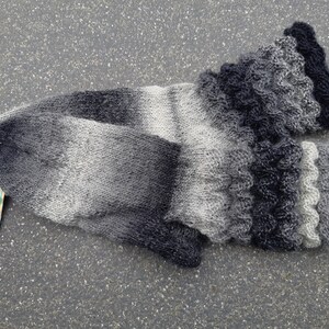 Könnte beinhalten: Ein Paar handgestrickte Socken in Grautönen, die von hell nach dunkel verlaufen. Die Socken haben einen gerüschten Rand und sind aus weichem, strukturiertem Garn gefertigt. Ein Etikett ist an einer Socke befestigt.