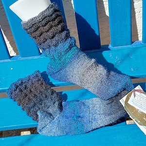 Pode incluir: Um par de meias feitas à mão em tons de azul, cinza e verde-azulado. As meias têm uma bainha texturizada e com babados e um padrão de cores gradiente. As meias são exibidas em uma superfície azul.