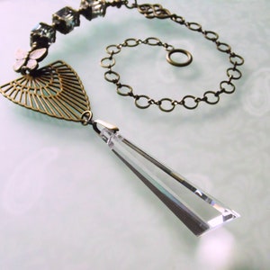 Könnte beinhalten: Eine goldene Kettenkette mit einem Kristallanhänger und einem dekorativen Metallanhänger. Der Anhänger ist fächerförmig und hat ein filigranes, kompliziertes Design.