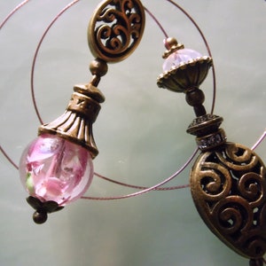 Könnte beinhalten: Nahaufnahme von zwei verzierten Ohrringen im antiken Stil. Jeder Ohrring hat ein bronzefarbenes Filigranmuster und eine rosa Glaskugel mit floralen Einschlüssen. Die Ohrringe sind durch einen dünnen, rosa Draht verbunden.