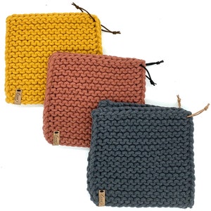 Könnte beinhalten: Drei gestrickte Stuhlkissen in verschiedenen Farben: Senfgelb, Terrakotta-Orange und Anthrazitgrau. Jedes Kissen hat ein braunes Leder-Etikett mit dem Text "Luna's Handmade Cushions".