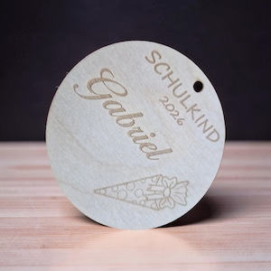 Puede incluir: Un adorno circular de madera clara con el nombre Gabriel, la palabra SCHULKIND, el a&ntilde;o 2026 y un grabado l&aacute;ser de un cono de regalo con un lazo. Tiene un peque&ntilde;o orificio circular en la parte superior para colgarlo.