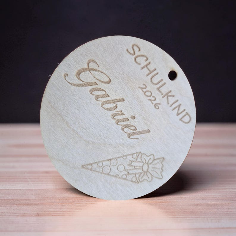 Puede incluir: Un adorno circular de madera clara con el nombre Gabriel, la palabra SCHULKIND, el a&ntilde;o 2026 y un grabado l&aacute;ser de un cono de regalo con un lazo. Tiene un peque&ntilde;o orificio circular en la parte superior para colgarlo.