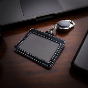 Puede incluir: Portatarjetas de identificación de cuero negro con costuras blancas y un carrete retráctil. El soporte tiene una ventana transparente para mostrar una tarjeta de identificación. El carrete tiene un clip de metal plateado y una carcasa de plástico negro. Sobre una superficie de madera.