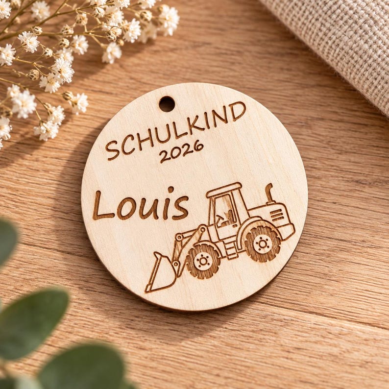 Puede incluir: Un adorno redondo de madera clara con un peque&ntilde;o orificio en la parte superior. La superficie est&aacute; grabada con l&aacute;ser con el texto SCHULKIND 2026, el nombre Louis y una ilustraci&oacute;n detallada de un tractor de construcci&oacute;n con pala cargadora.