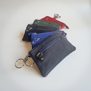 Könnte beinhalten: Eine Auswahl an kleinen Leder-Münzbörsen in verschiedenen Farben: Marineblau, Königsblau, Schwarz, Grün und Rot. Jede Börse hat einen Reißverschluss und einen silbernen Schlüsselring. Die Börsen sind leicht überlappend fächerförmig angeordnet.