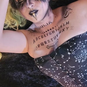 May include: A black corset with a white constellation pattern and a black and white Ouija board printed on the front. The model is wearing the corset and has a black and white face paint design.