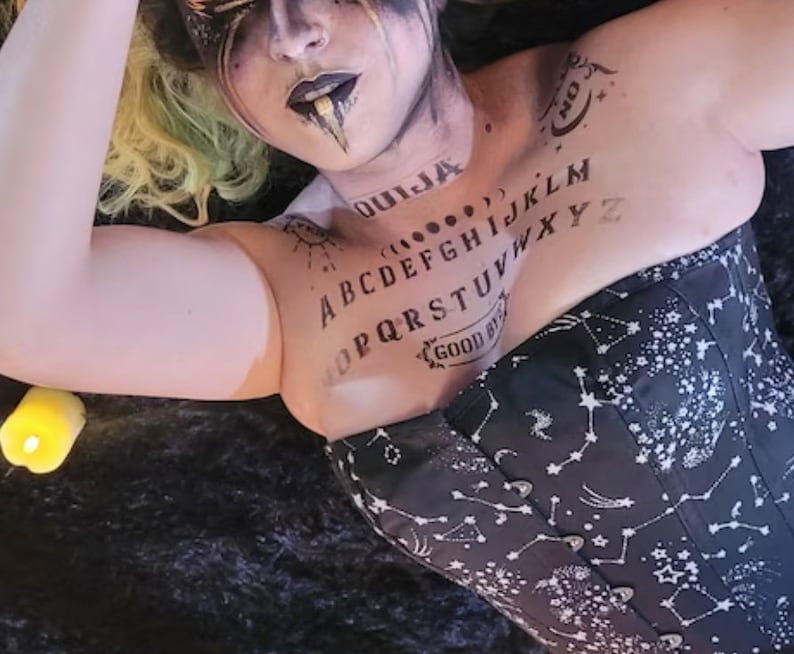 May include: A black corset with a white constellation pattern and a black and white Ouija board printed on the front. The model is wearing the corset and has a black and white face paint design.