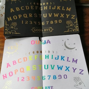 May include: Two open notebooks with a Ouija board printed on each page. The top notebook has a black background with gold lettering and symbols. The bottom notebook has a white background with colorful lettering and symbols. The word "OUIJA" is printed in rainbow colors at the top of the bottom notebook.